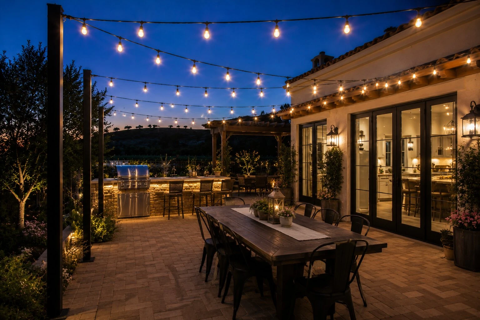 Spanish revival home at deep-blue dusk with warm bistro string lights on dedicated black steel posts, a paver herringbone patio, a farmhouse dining table in the foreground with lanterns and a centerpiece, an outdoor kitchen with bar stools in the mid-ground, vineyard rows behind