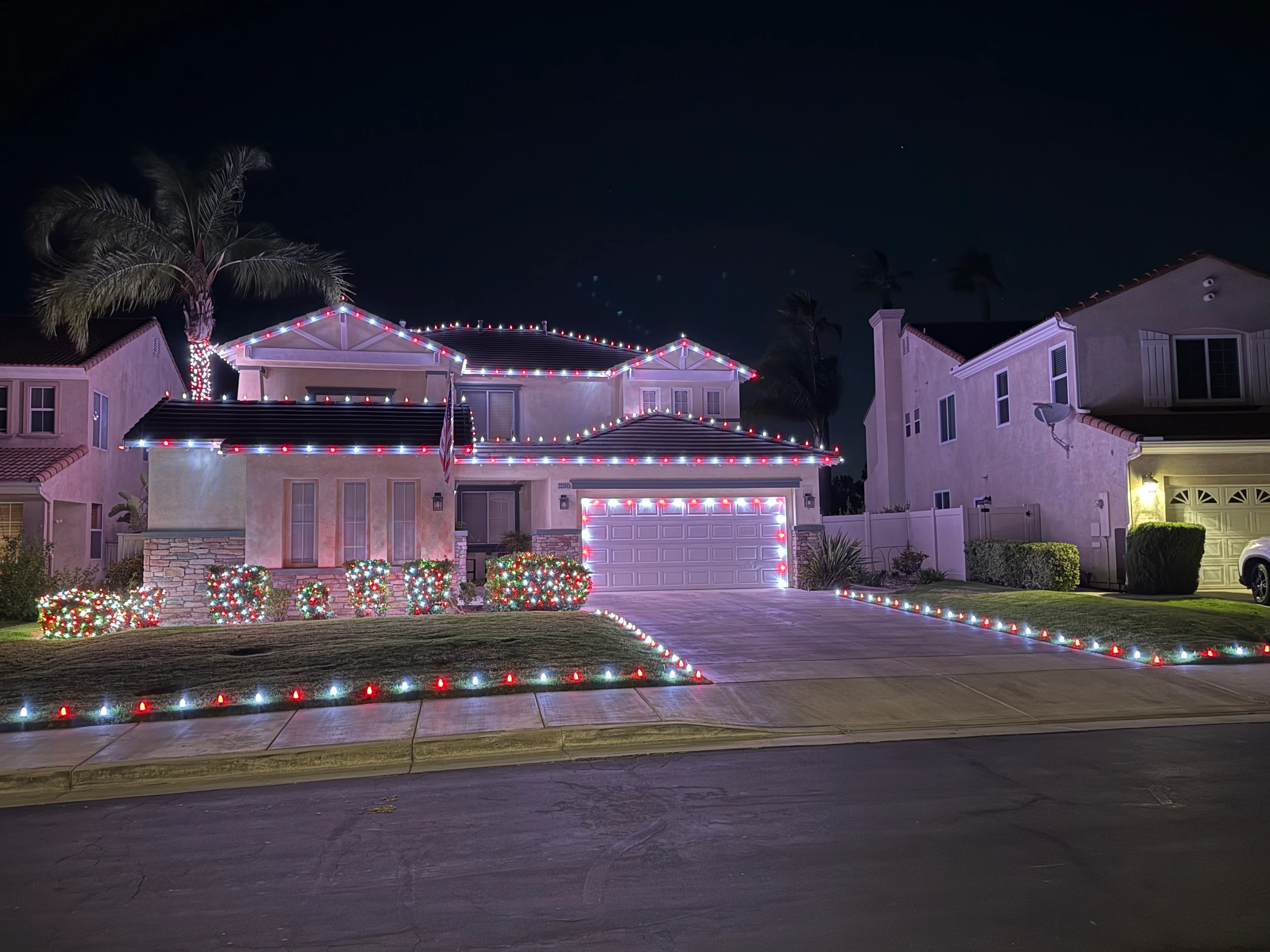 Recent Christmas project: single-story residence at night with multicolor C9 Christmas lights tracing every gable, ridge, and garage door, landscape mounds outlined in red and white at the curb, one front-yard palm wrapped warm-white at the entry for contrast