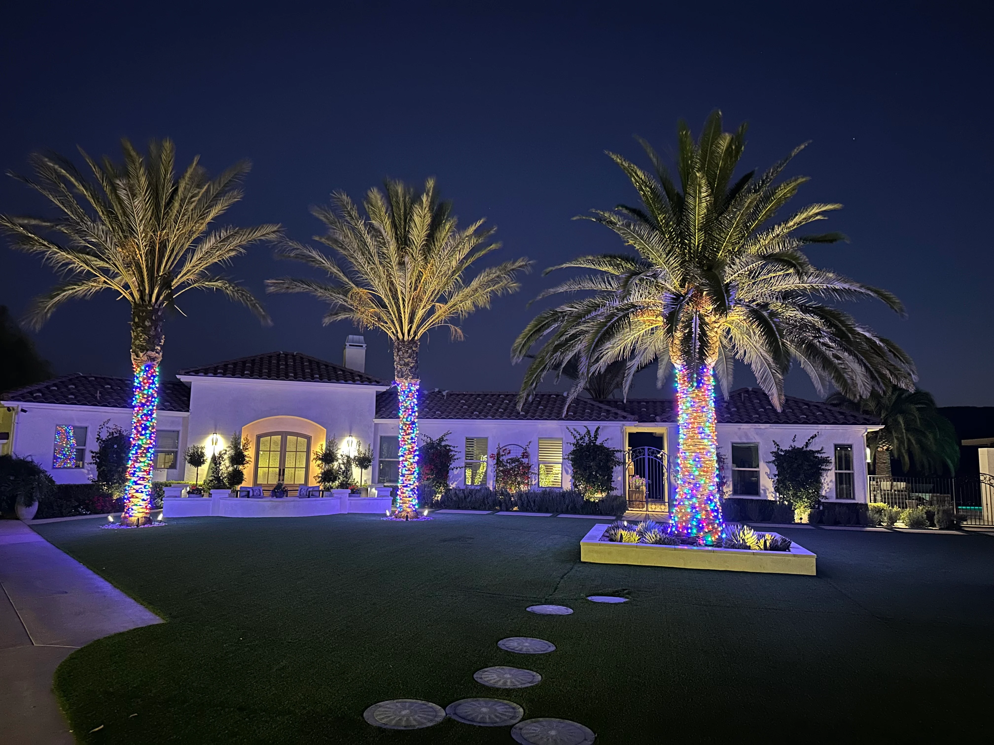Single-story Spanish revival home at dusk with three front-yard palms wrapped in multicolor Christmas lights, terracotta tile roof, stone walkway