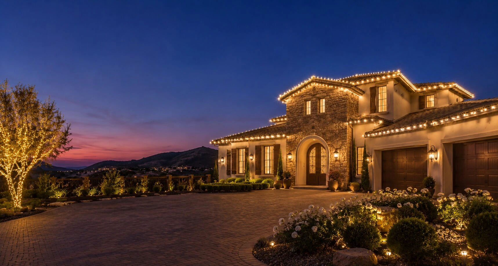 Two-story Tuscan stone estate at dusk with warm-white Christmas C9 lights traced along every gable and ridge, a large wrapped tree at left, paver driveway, pink and blue sunset behind the mountains