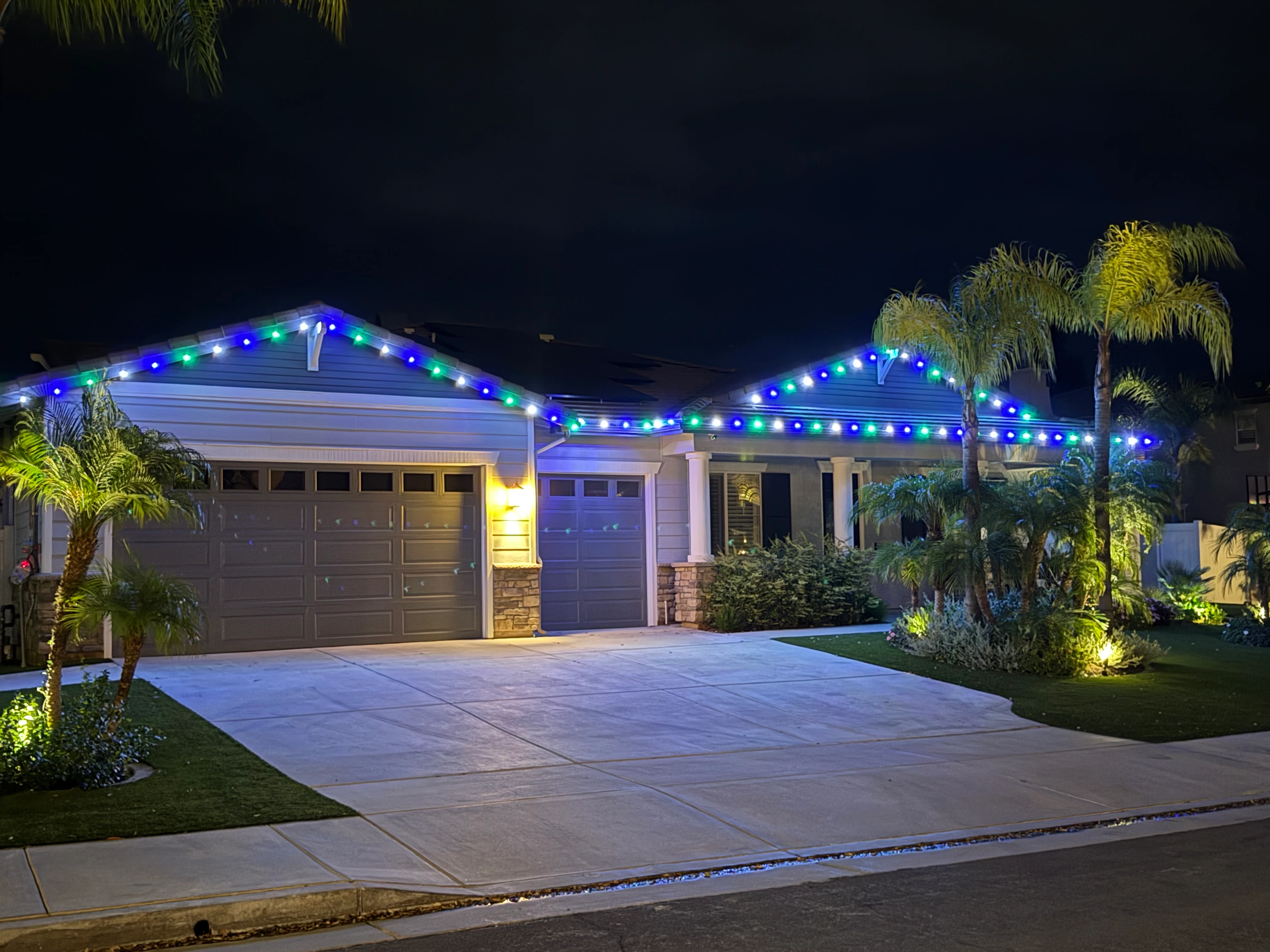Single-story Modern Craftsman home at dusk with blue and green multicolor C9 lights traced along two gables, two front-yard palms wrapped warm-white