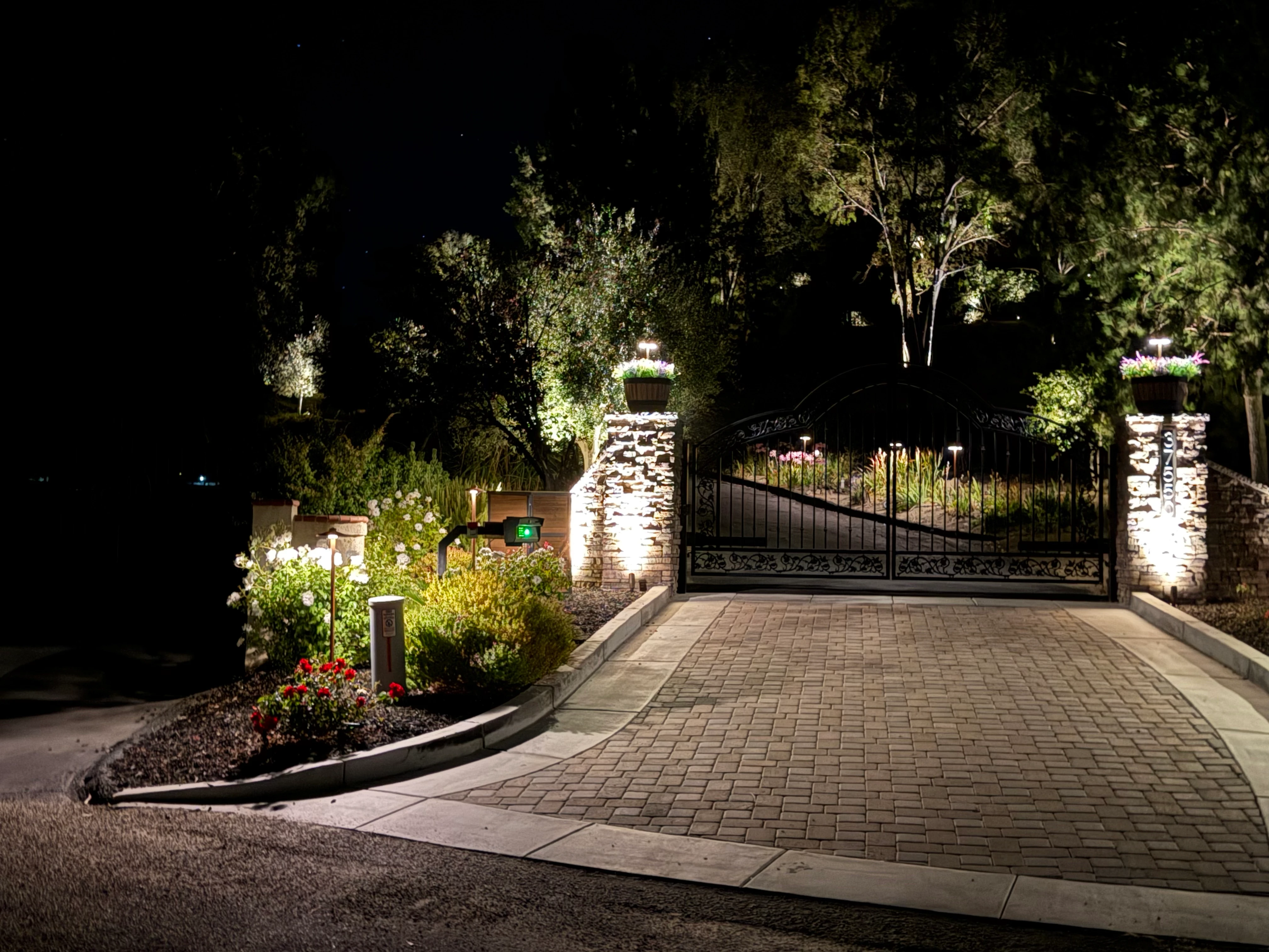 Estate gate at dusk with stone pillars and iron gate, urn planters with warm-white wrapped olive trees on top, paver driveway in foreground