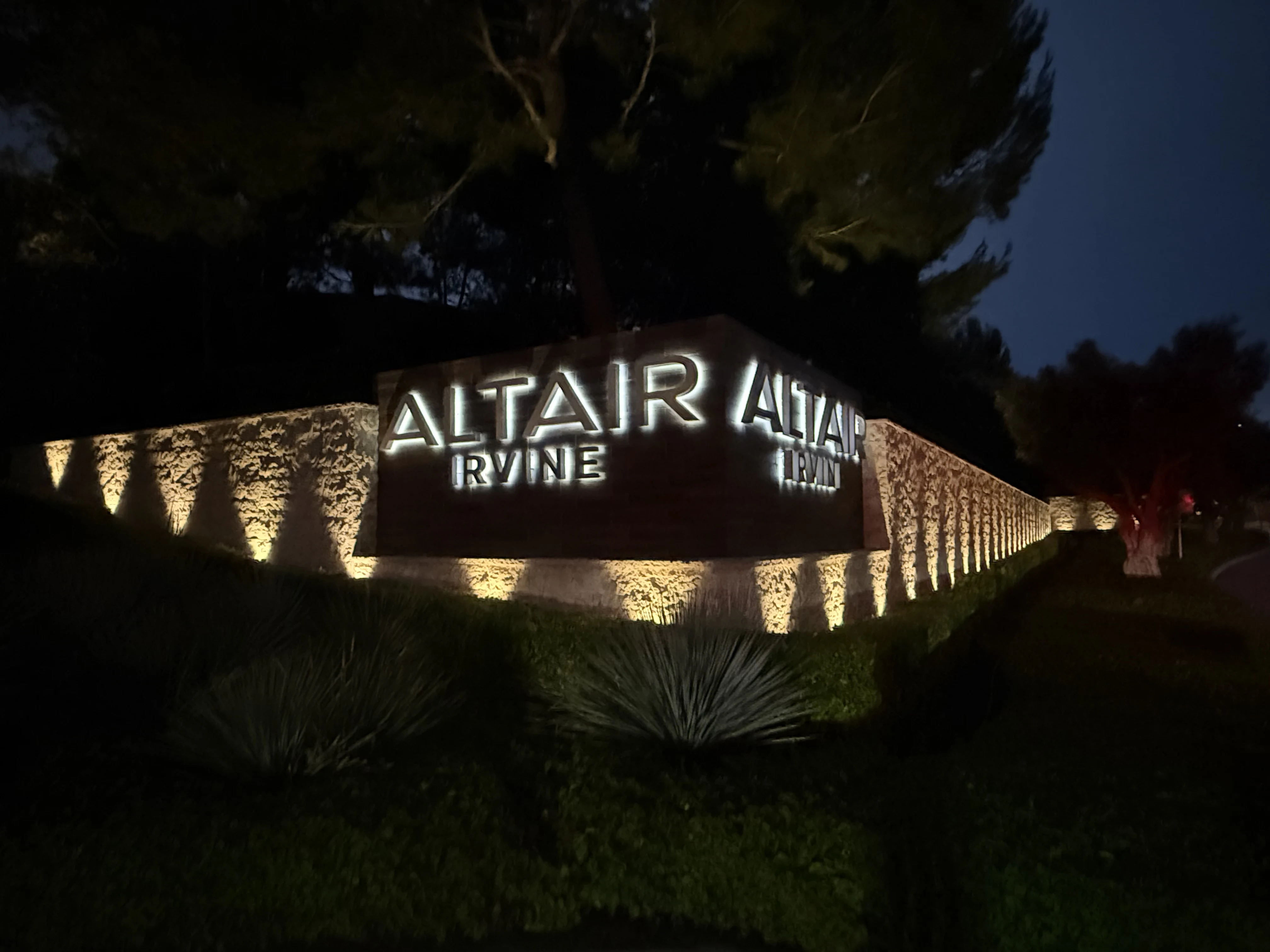 Backlit halo cursive lettering on a stacked-stone HOA monument at night, uplit from the base with brass fixtures, tall pine canopy uplit behind, the Altair Irvine community entry signage