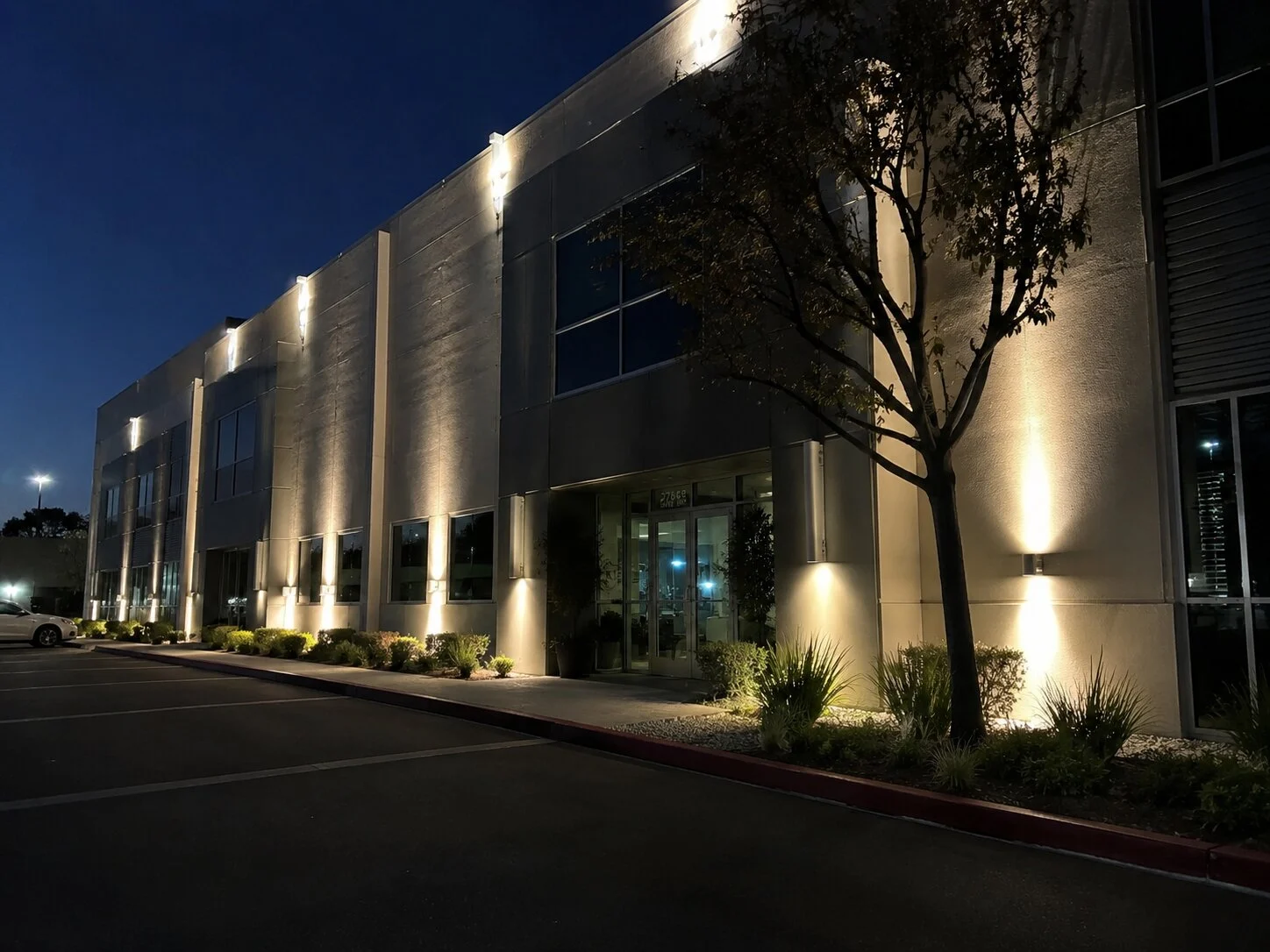 Two-story Class-A office building at dusk with up-and-down wall pack fixtures along the stucco facade, small trees uplit in the landscape beds, clean parking lot and address "27563" visible
