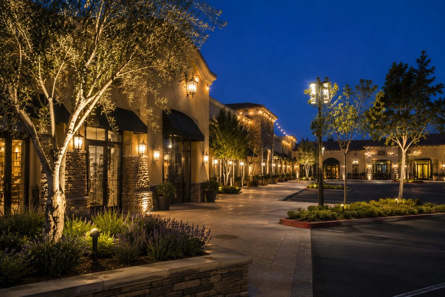 Spanish revival retail main street at dusk with warm downlight lanterns mounted on every facade, small olive trees uplit in stacked-stone planters, lit street lamps along the boulevard