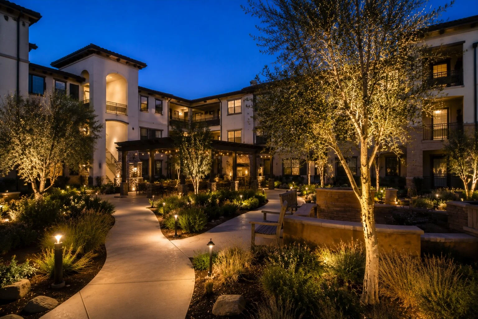 Spanish revival multifamily residential complex at dusk with path lights along a curved walkway, tree uplighting on white-bark olives, multiple lit balconies and arched entries visible across the courtyard
