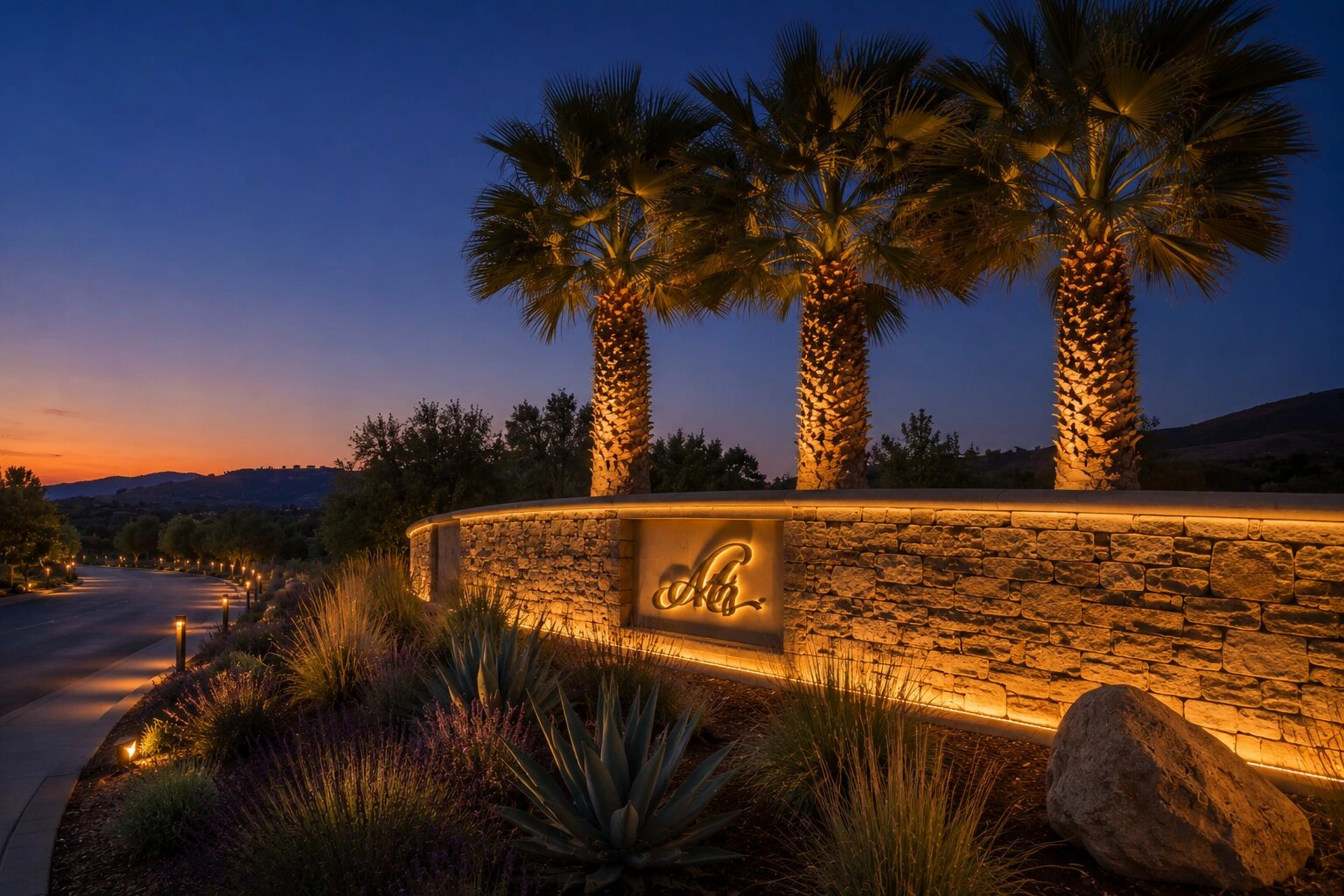 Three uplit palm trees flanking a stacked-stone HOA entry monument with a cursive logo sign and warm desert sunset behind, native landscaping and bollard path lights along the entry road