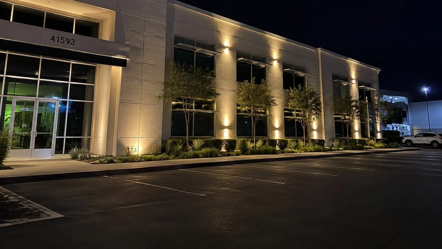 Two-story Class-A office building "41593" at dusk with up-and-down wall pack fixtures spaced evenly along the stucco facade, small trees uplit at the base of the building, paver landscape strip in the foreground