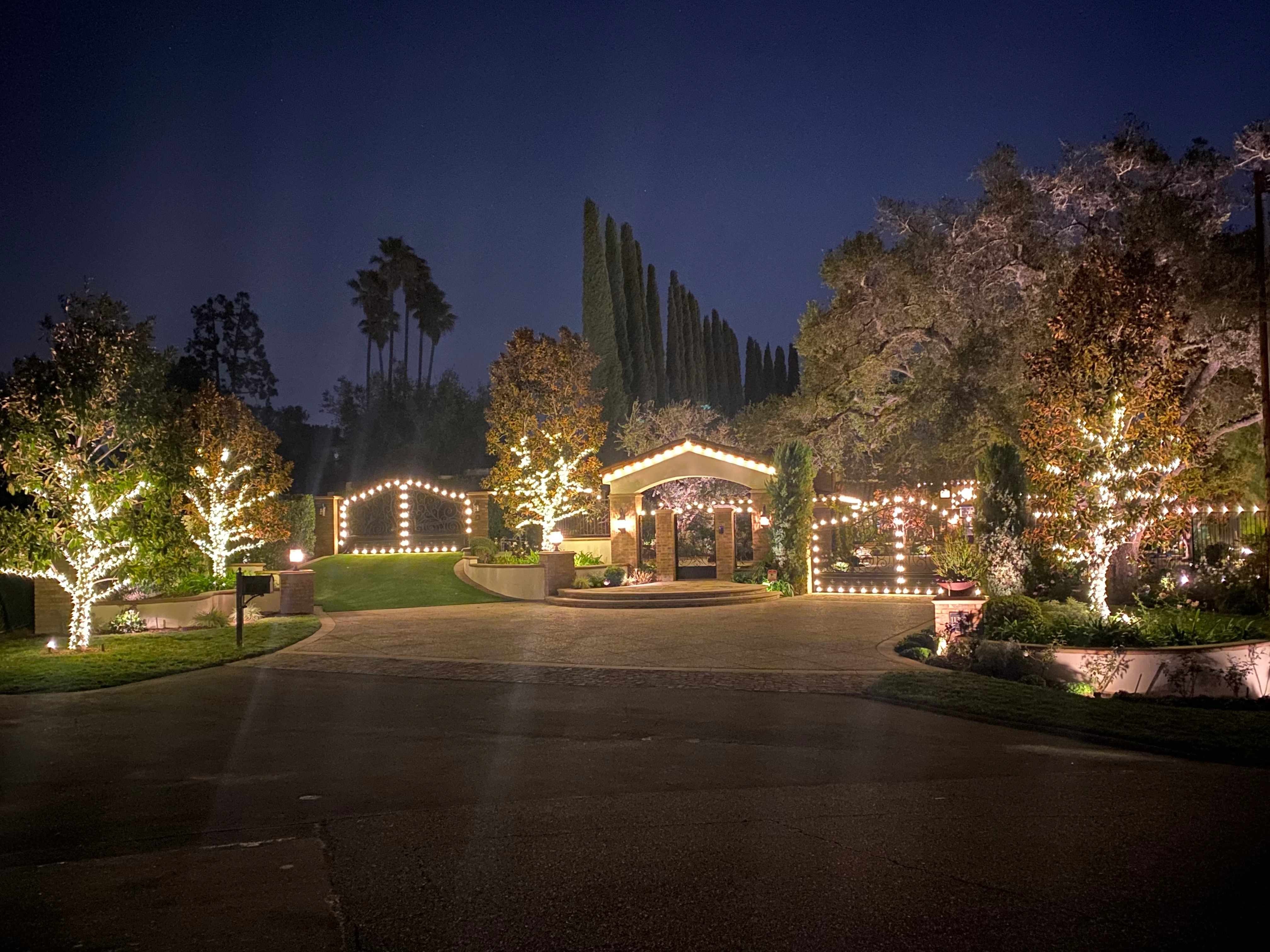 Wide Mediterranean estate driveway outlined in warm-white Christmas lighting at dusk, with cypress and palm wraps along the entry, Temecula