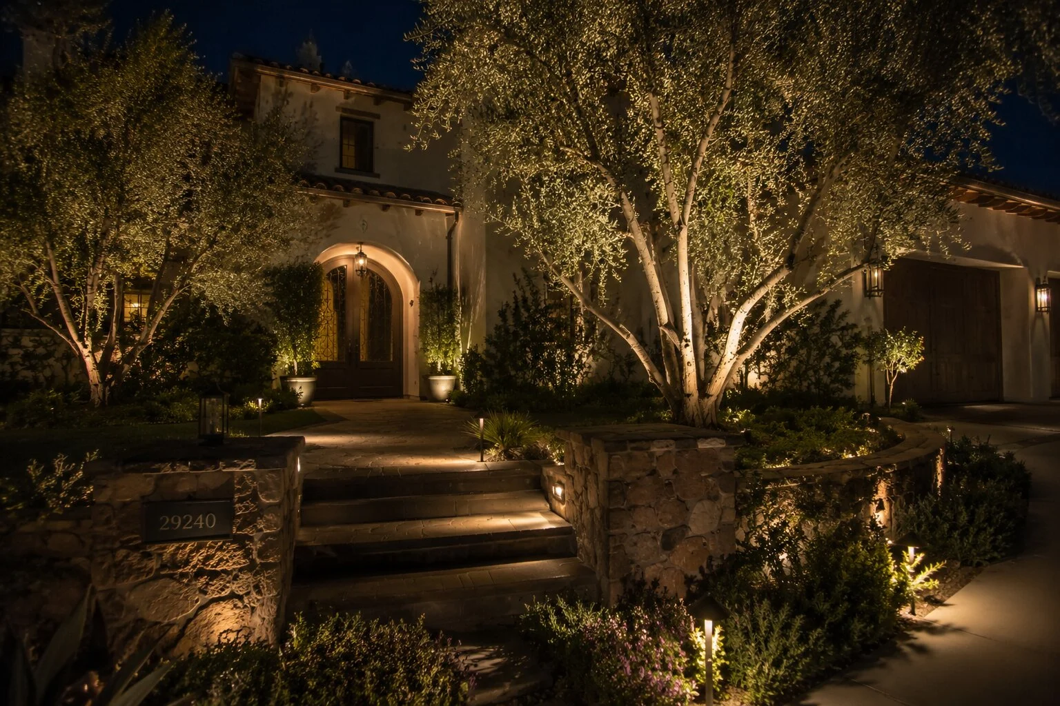 Spanish-style entry at dusk with arched front door, uplit olive trees, low path lights, and a stone mailbox set into a planter wall