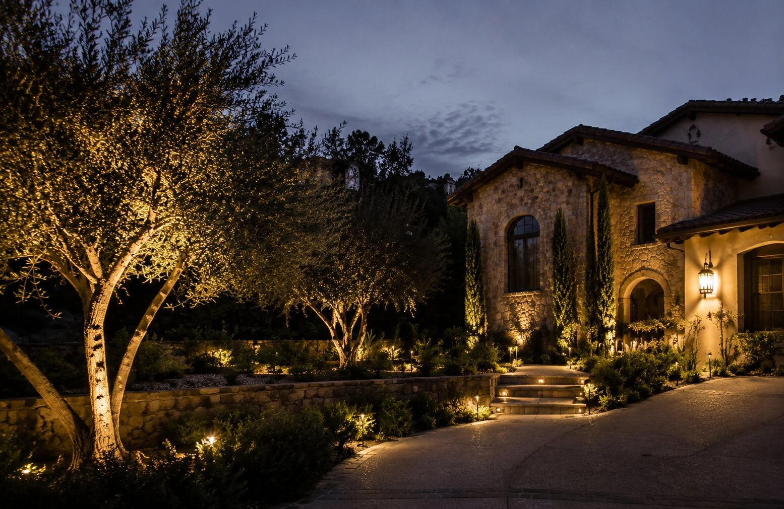 Stone Tuscan-style estate at dusk with multiple olive trees uplit and warm windows glowing, Temecula Valley