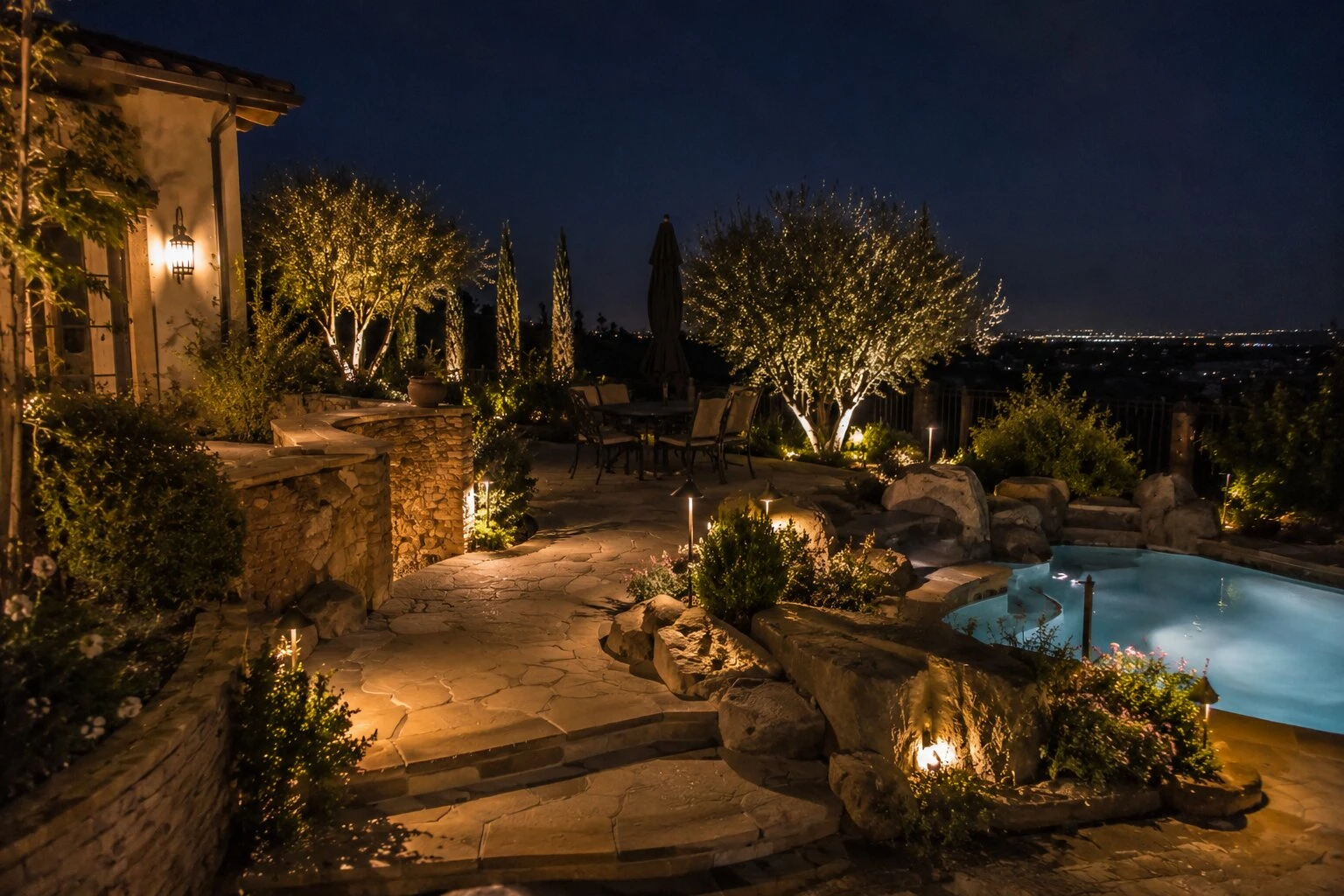 Hilltop pool deck at dusk with flagstone patio, uplit palm and olive trees, and the lights of the valley in the distance