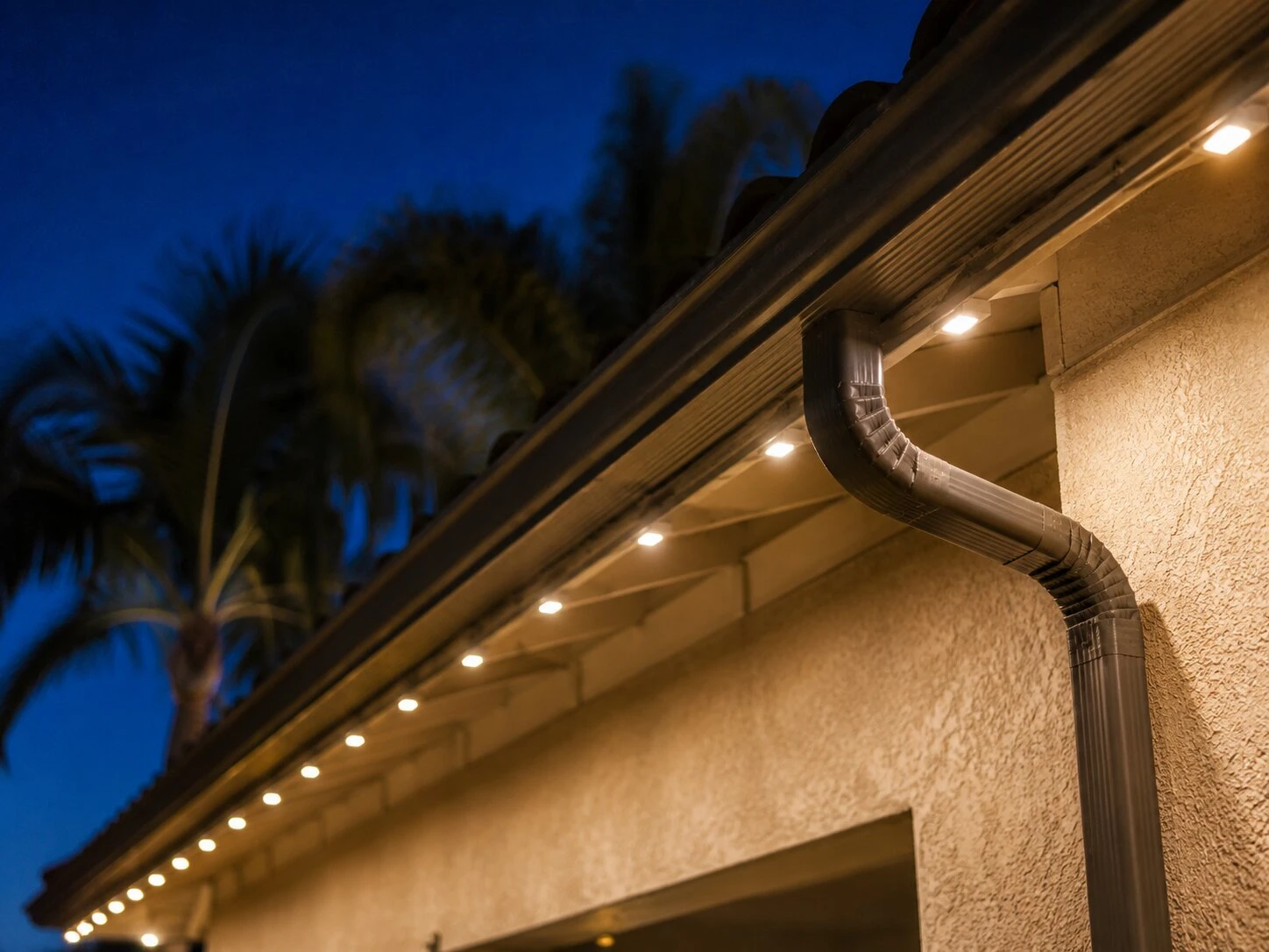 Close-up of warm-white eave-mounted permanent LED dot lights running under the soffit of a stucco home at dusk, deep-blue sky and palm fronds behind, slim trim channel visible at the fascia
