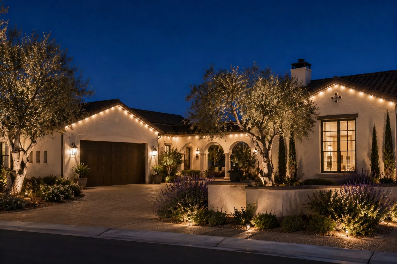 Single-story Spanish-style home in the Temecula Valley at deep-blue dusk with warm-white permanent LED lights running under every eave and gable, mature olive trees uplit from the ground, paver driveway and lavender-lined planter beds