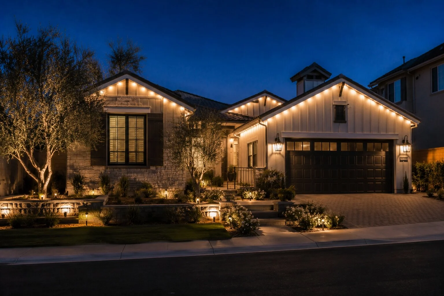 Modern farmhouse home in stone and board-and-batten at dusk with warm-white permanent LED lights running under every gable and ridge, paver driveway, olive tree uplit at left