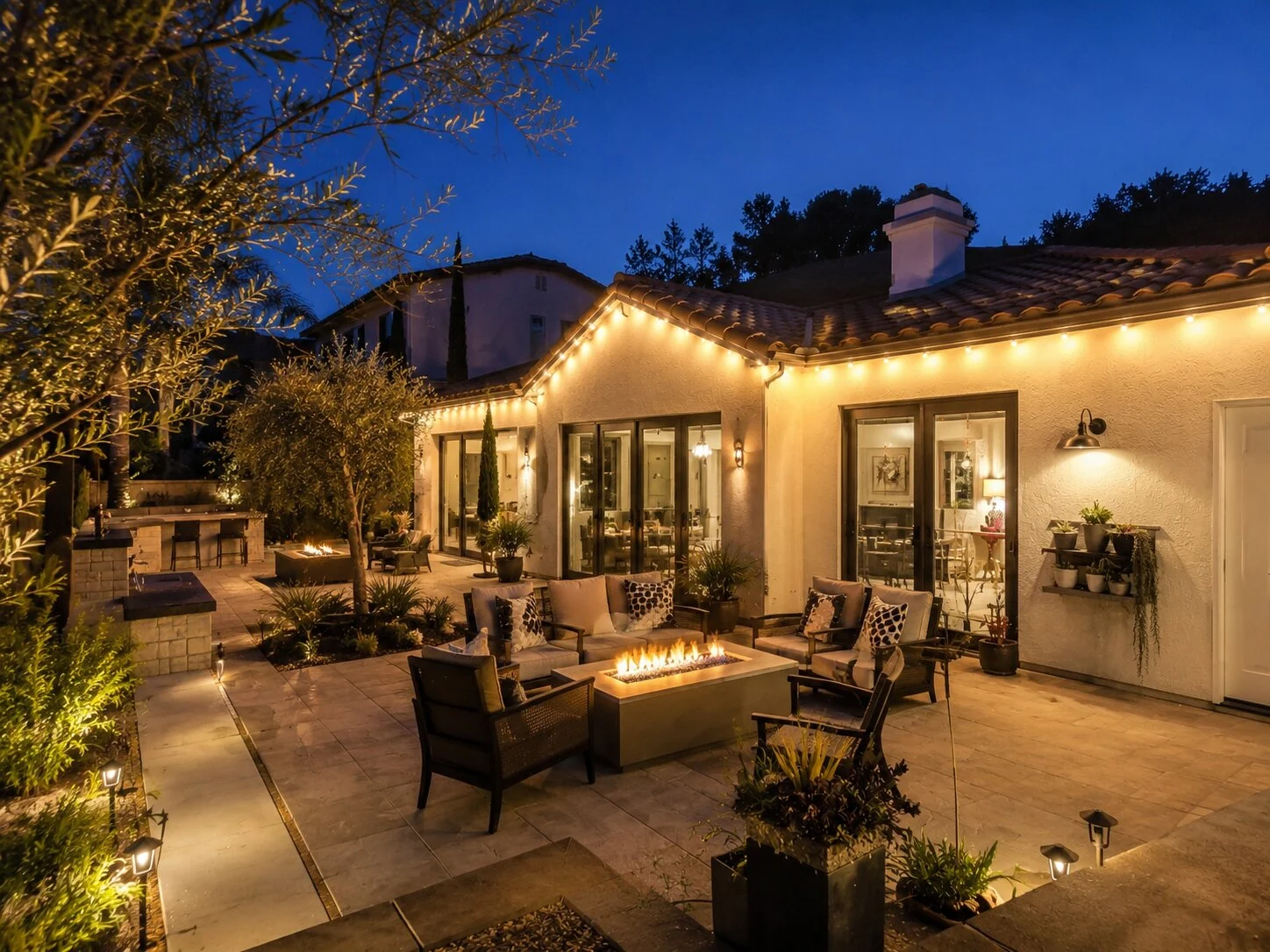 Spanish patio at dusk with warm-white permanent LED lights along the eave, a low gas firepit with seating around it, olive trees uplit at left, dining and lounging beyond