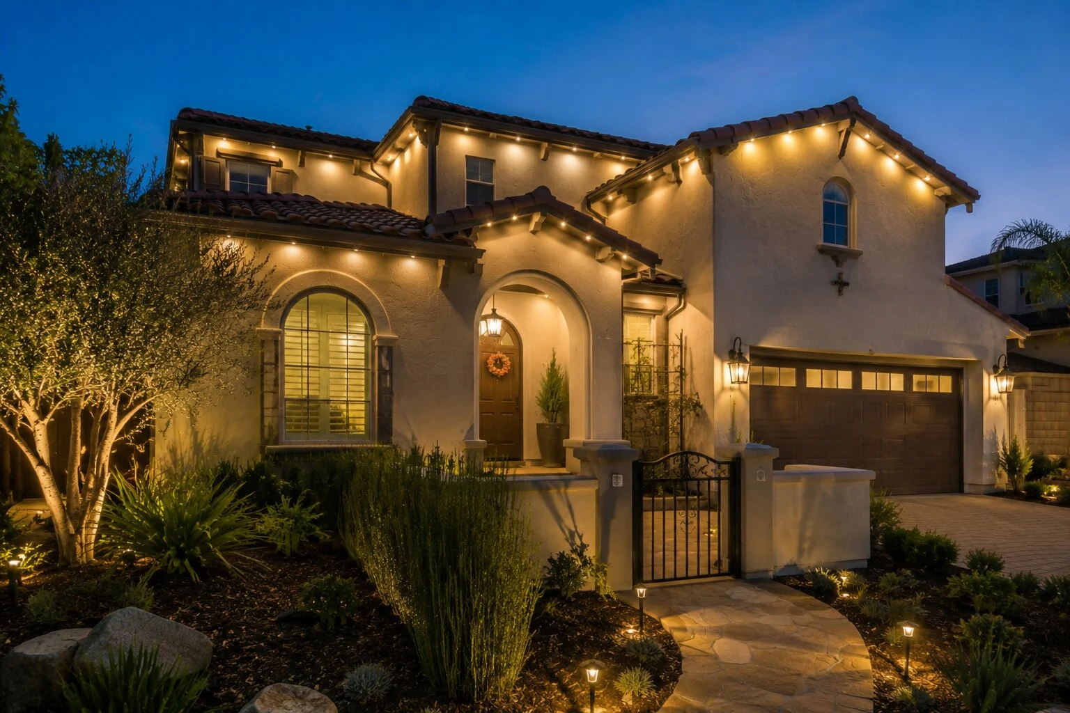 Two-story Spanish home at dusk with warm-white permanent LED lights along the eave, arched front windows and entry, low path lights along the landscape beds and front walk