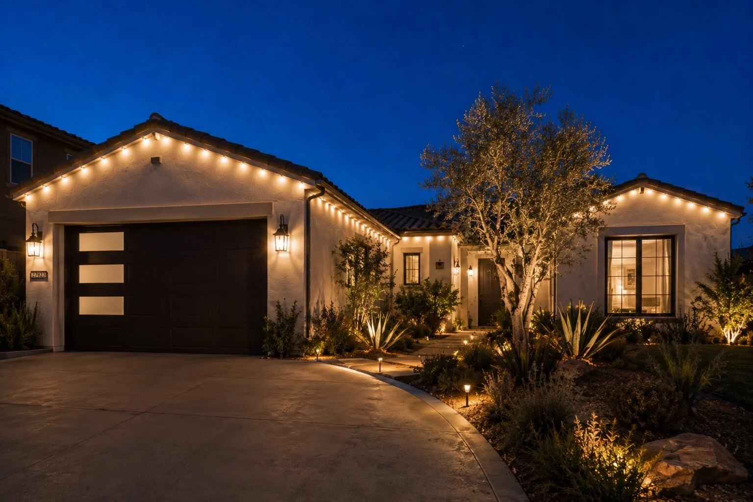 Single-story modern Spanish home at dusk with a dark windowed garage door and warm-white permanent LED lights along the eave, mature olive tree uplit from the ground, agave path-light specimens in the bed