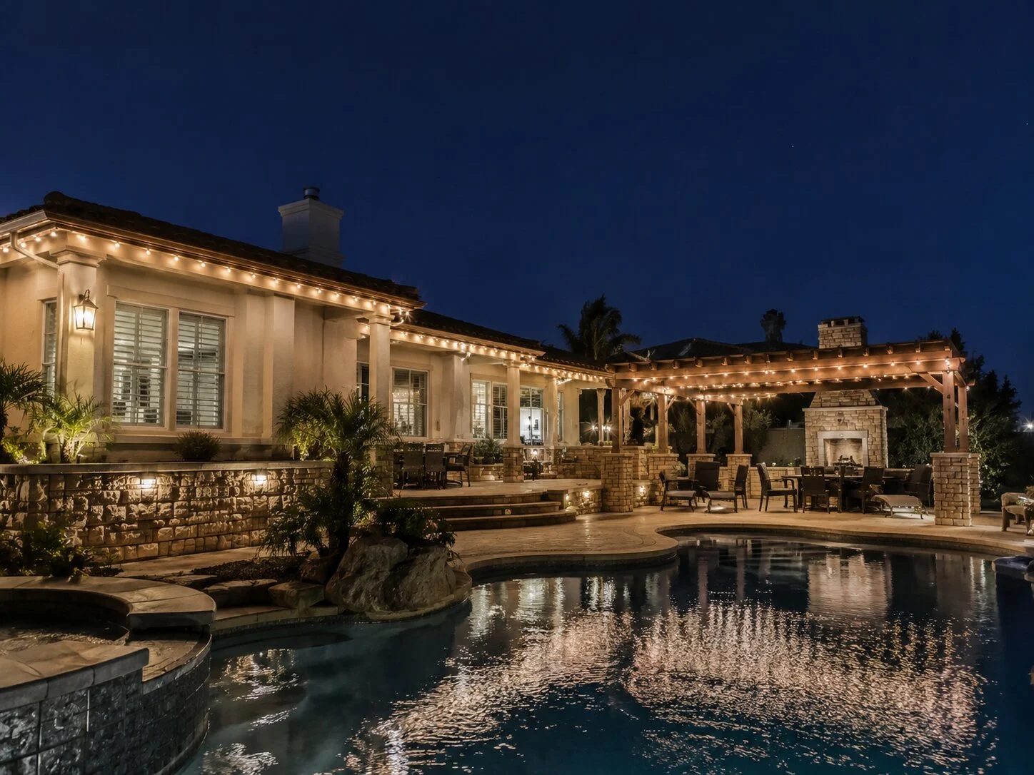 Large Spanish home with covered pergola and outdoor fireplace at dusk, pool reflection in the foreground, warm-white permanent eave lighting throughout