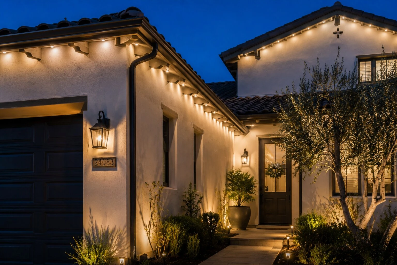 Spanish-style front entry at dusk with warm-white permanent LED lights running under the eave, gas-style entry lanterns flanking the front door, olive tree at right, address numbers visible on the stucco