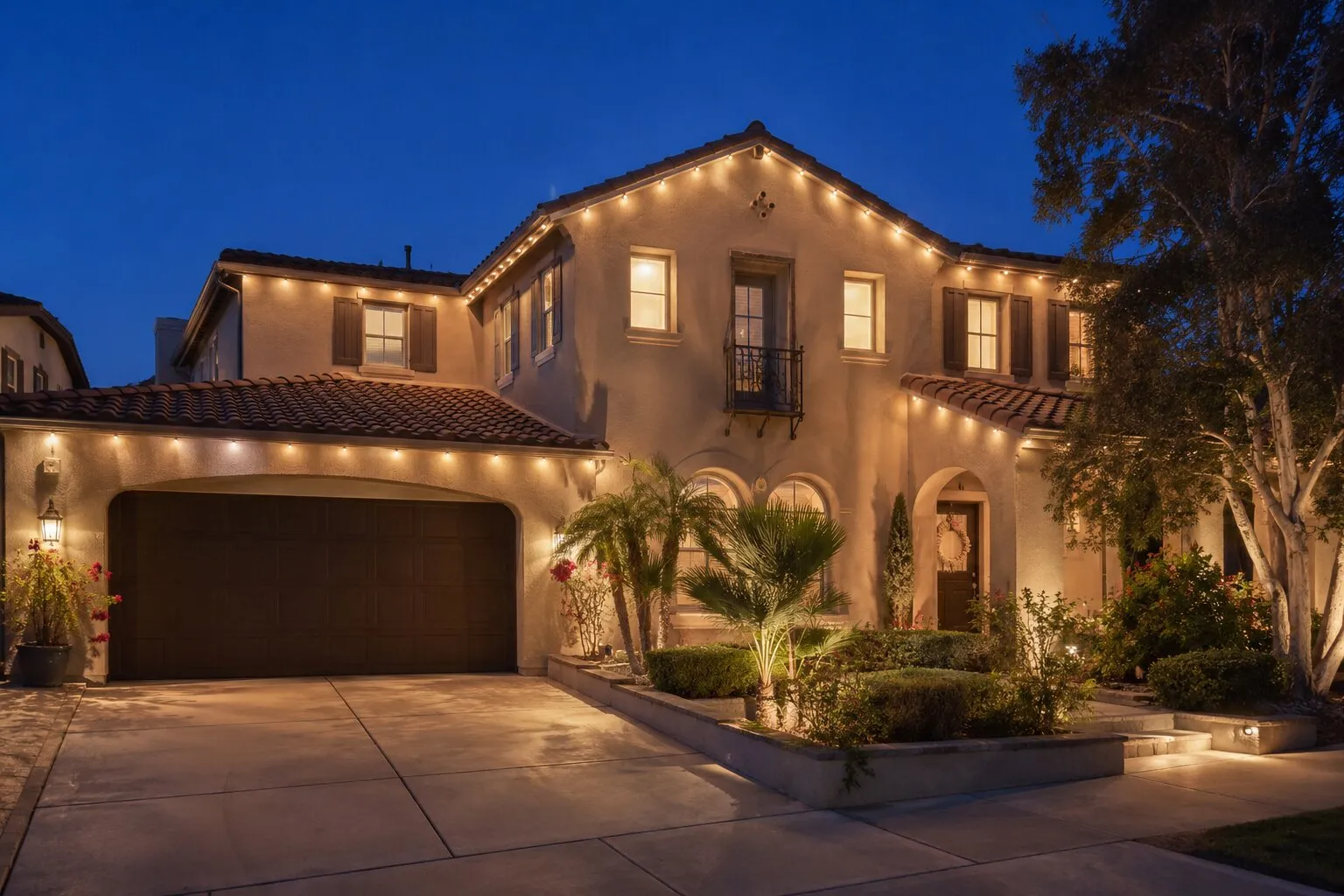 Two-story Spanish revival home at deep-blue dusk with warm-white permanent LED lights running under every eave and gable, terracotta tile roof, a small palm and roses by the entry, paver driveway