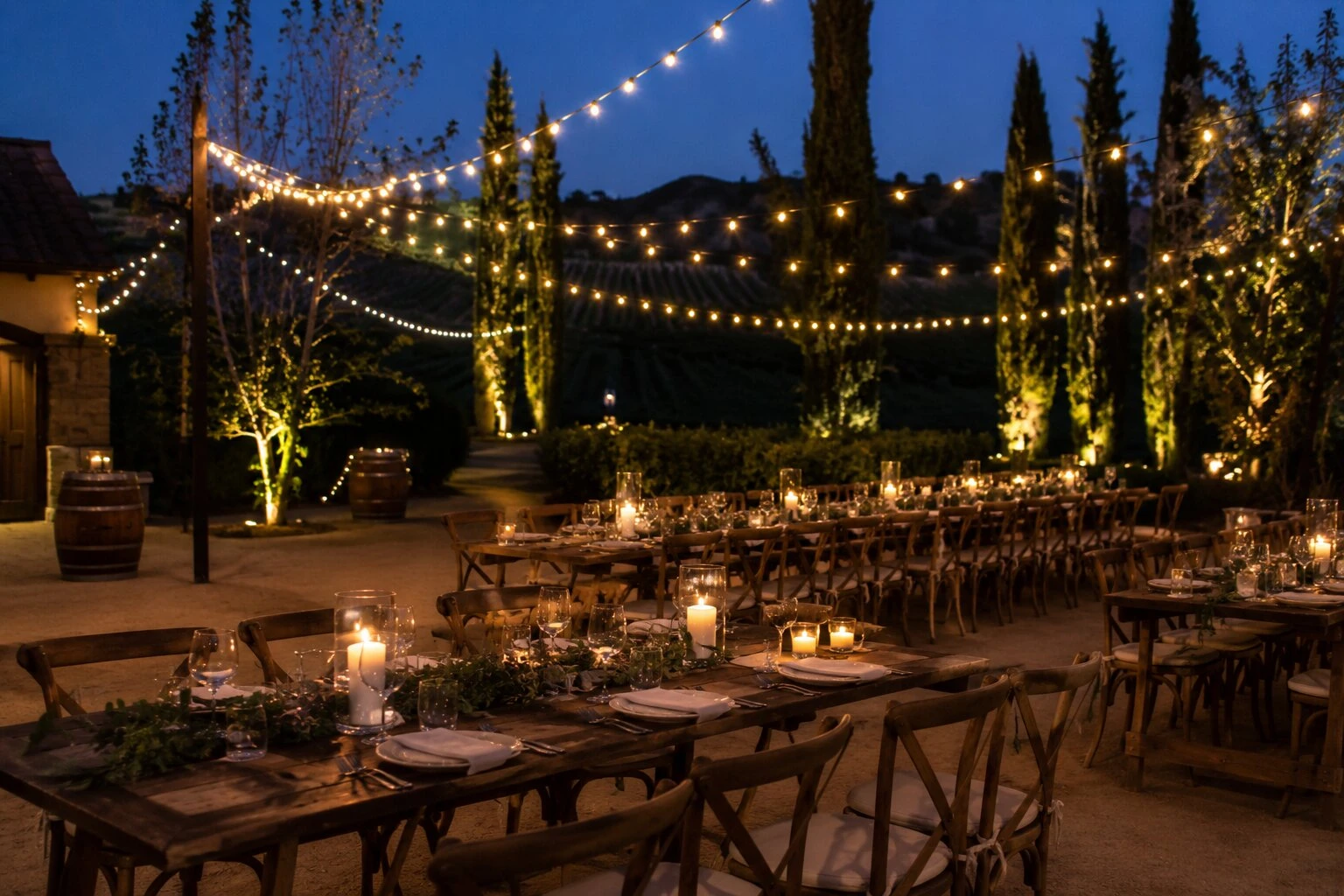 Close detail of a vineyard reception table at dusk with hurricane-glass candles glowing on a wooden farmhouse table, tall cypress columns flanking, warm bistro string lights overhead, wine barrels at the edge of the frame