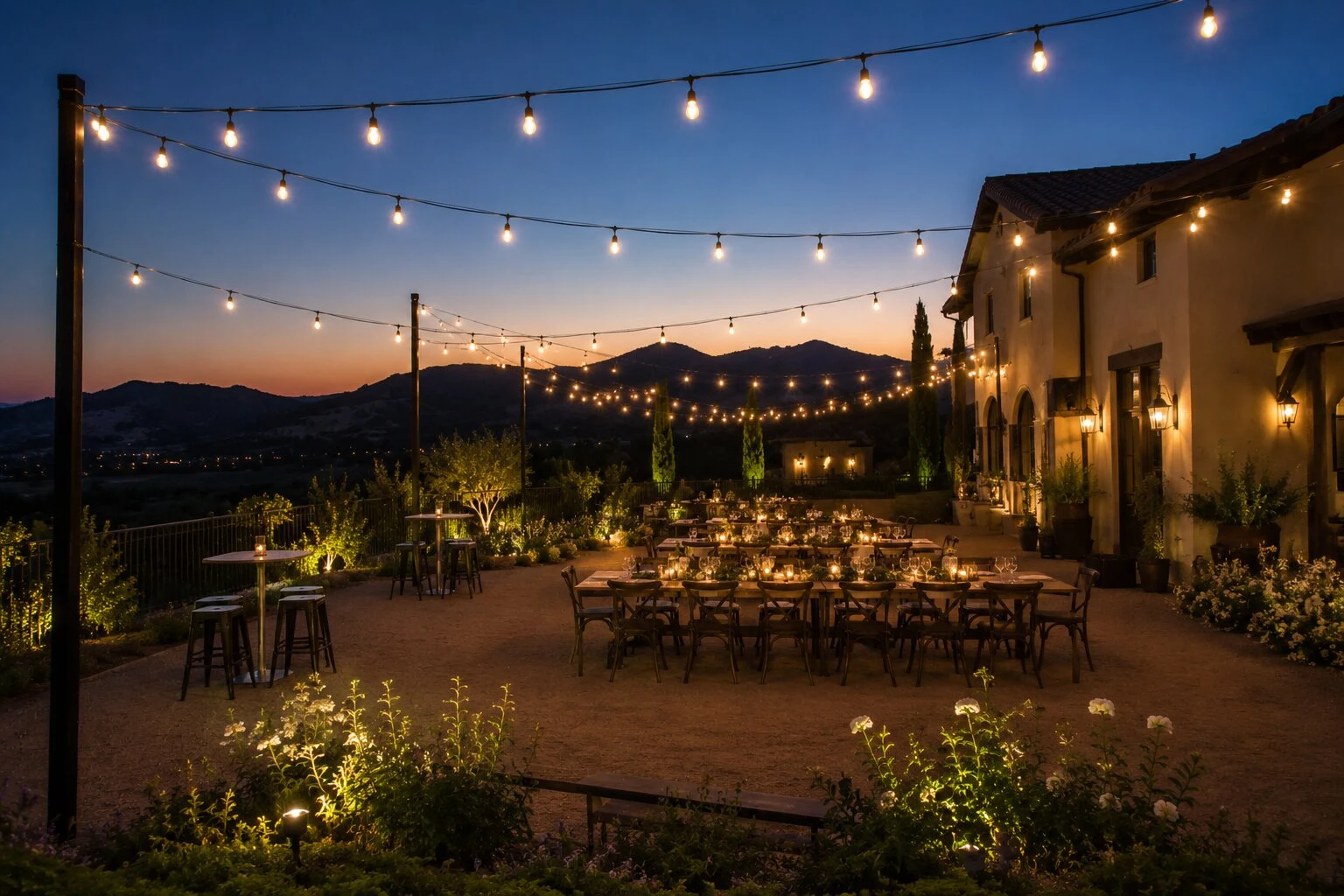 Mountainside Tuscan terrace reception at sunset with cascading warm bistro string lights overhead, round candle-lit tables on a paver patio, garden flowers in the foreground, valley views and a silhouette mountain ridge under a pink and orange sunset sky