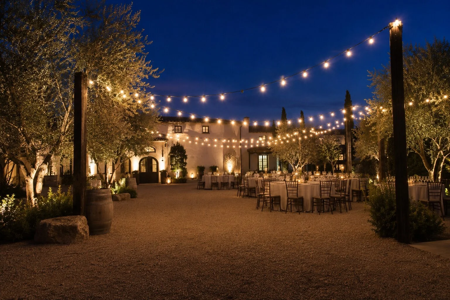 Spanish revival courtyard reception at dusk with round farmhouse tables and chiavari chairs on a gravel patio, warm bistro string lights crisscrossing overhead between olive trees and an arched stone entry