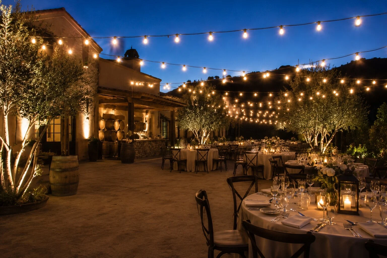 Tuscan-style courtyard dinner at dusk with round tables lit by hurricane candles and chiavari chairs, warm bistro string lights overhead, wine barrels by the bar, mountain hillside in the distance