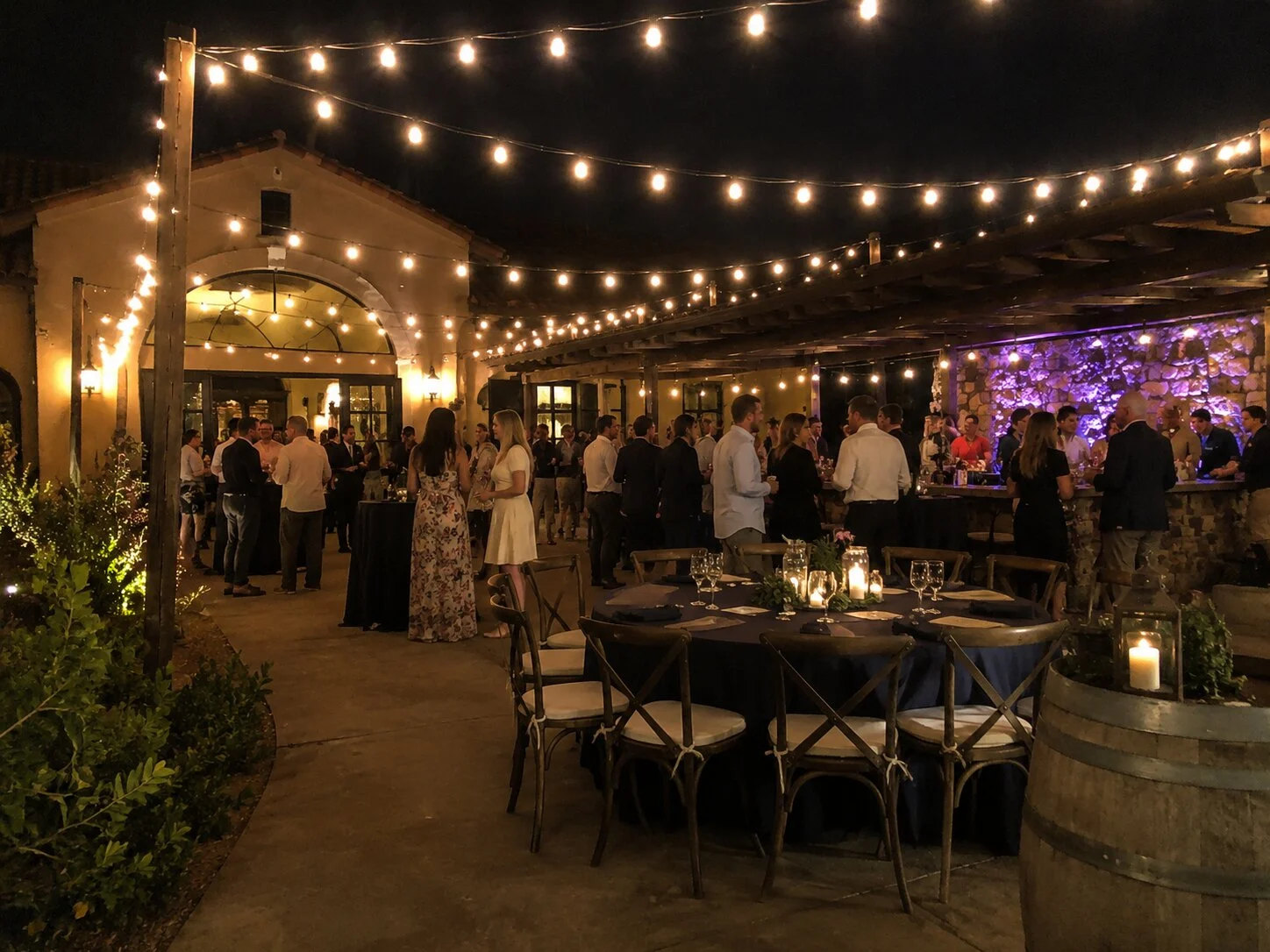 Cocktail reception with mingling guests at night, warm bistro string lights overhead, Spanish revival archway and a stone bar with purple accent uplighting on the back wall, dressed cocktail tables in the foreground