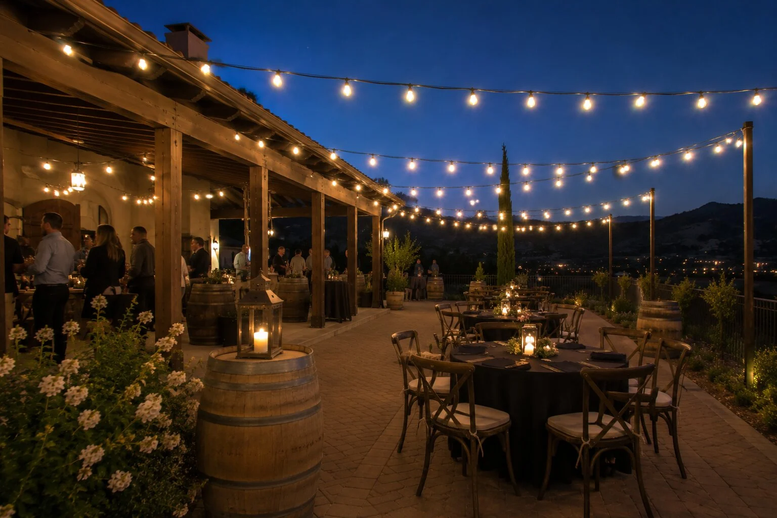 Wine-country terrace cocktail reception at dusk with mingling guests on a paver patio, warm bistro string lights overhead, wine-barrel cocktail tables with lanterns, an open pergola and a mountain valley view in the distance