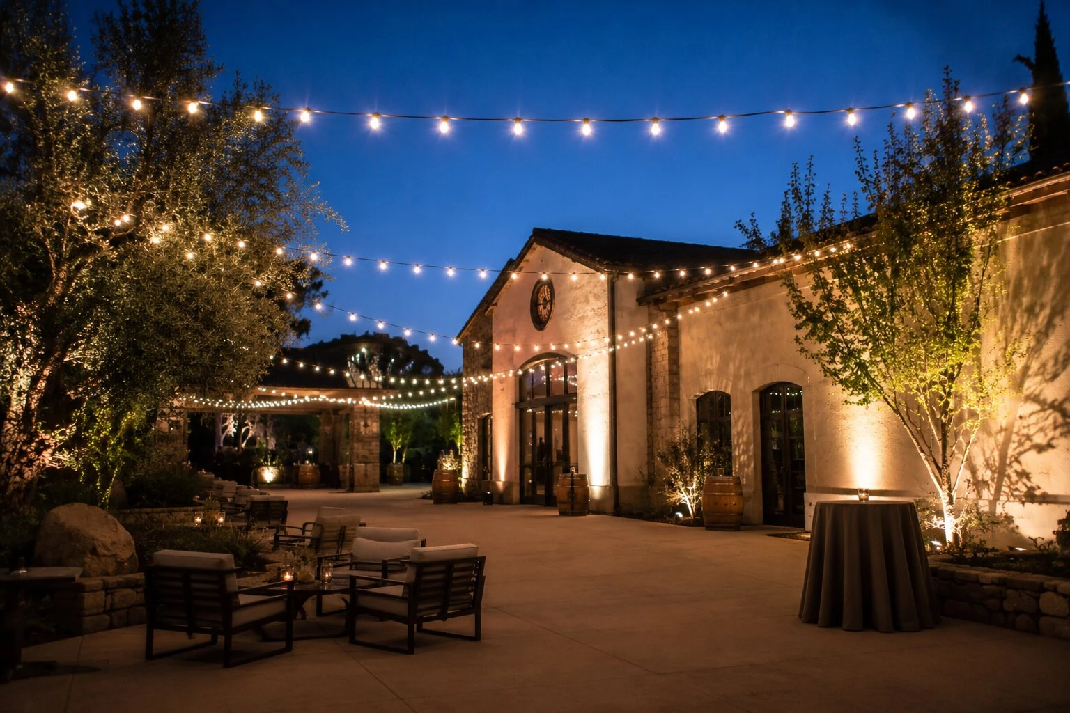 Spanish revival winery courtyard at dusk with warm bistro string lights overhead, an arched stone doorway with warm interior glow, wine barrels and lounge seating on a paver patio, olive trees flanking