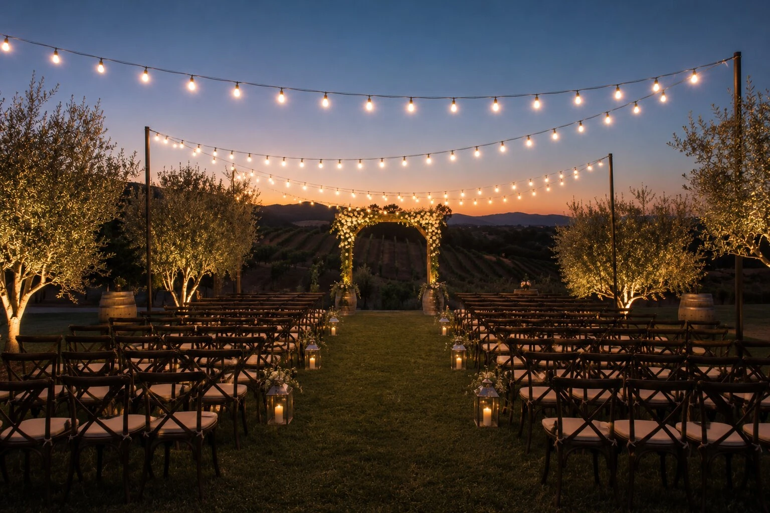 Outdoor vineyard wedding ceremony at sunset with rows of farmhouse chairs lined along a grass aisle, a floral arch at the head of the aisle, warm bistro string lights overhead, hanging lanterns along the aisle, vineyard rows and a mountain ridge in the distance