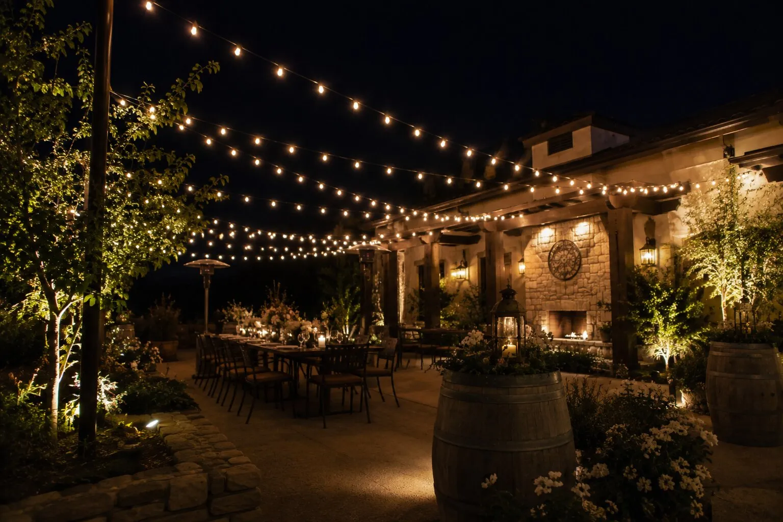 Intimate dinner setup under a wooden pergola at night with a stacked-stone fireplace at center, hurricane candles glowing along a long farmhouse table, warm bistro string lights running overhead, wine barrels and lanterns in the foreground